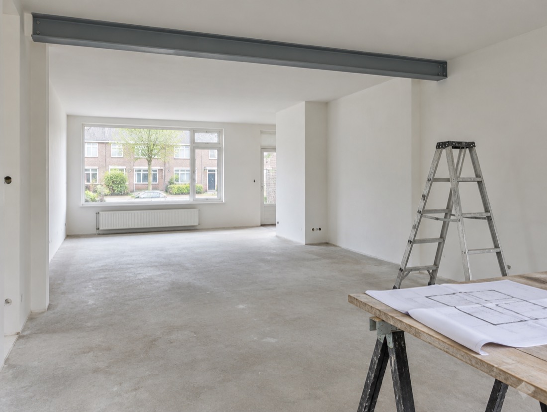 Een woonkamer waarin een wand is weggehaald, met een stalen balk in het plafond en een tekening op tafel